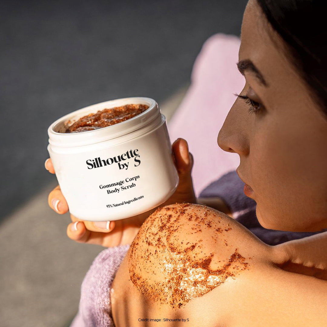 Femme regardant par-dessus son épaule gauche, tenant un pot de gommage corps Silhouette by S dans sa main droite, avec une touche de produit appliquée sur son épaule. Ce gommage exfoliant à la bave d’escargot élimine les cellules mortes, régénère et hydrate la peau pour un toucher doux et une texture lisse