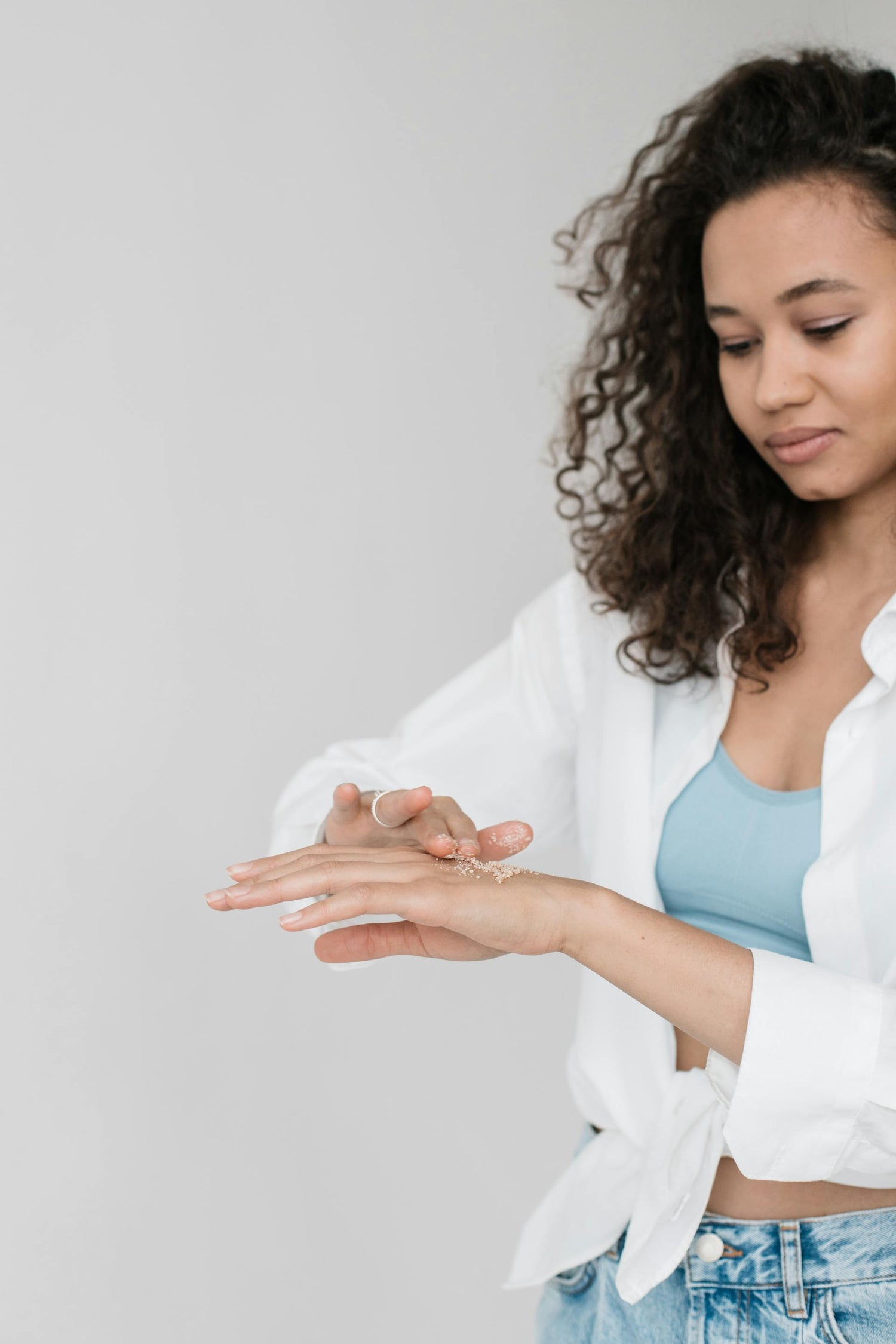 Jeune femme créole appliquant délicatement un gommage naturel sur sa main, exfoliant la peau pour éliminer les cellules mortes et améliorer la douceur et la texture. Ce soin exfoliant aux ingrédients naturels prépare la peau à mieux absorber les soins hydratants, tout en laissant un toucher soyeux et revitalisé. Un geste essentiel pour une routine beauté efficace qui allie bien-être et soin