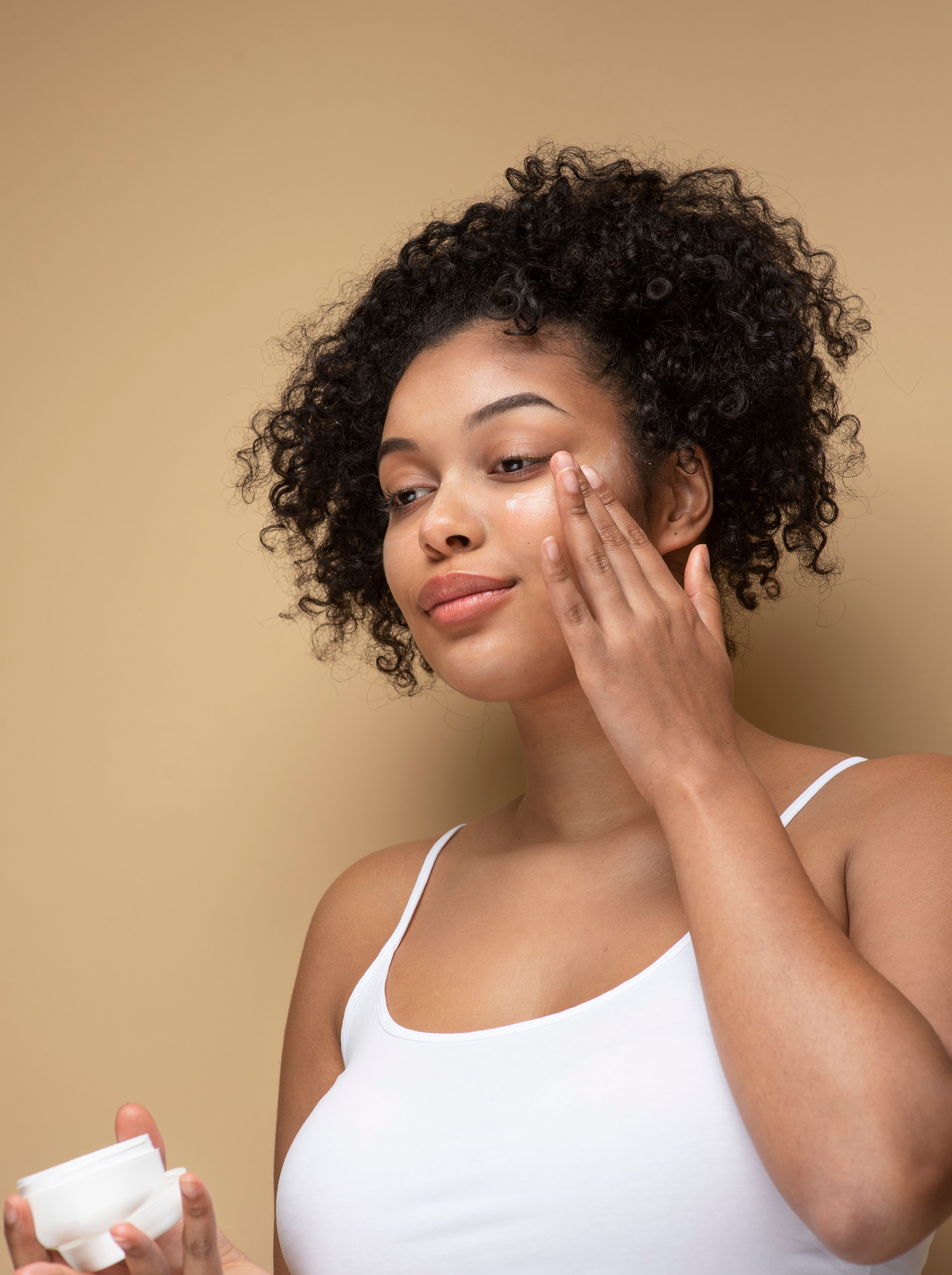 Femme métisse appliquant une crème sur son visage, un soin riche en bave d’escargot, acide hyaluronique et collagène. Cette crème tonifie, lisse et rajeunit la peau en réduisant rides et ridules, tout en illuminant le teint. Sa texture légère pénètre rapidement sans laisser de film gras, offrant un effet liftant visible dès les premières utilisations. Convient à tous types de peau, formulée avec 99% d’ingrédients naturels et testée sous contrôle dermatologique